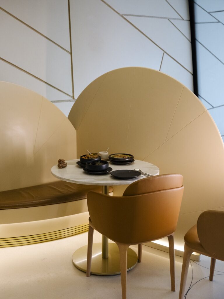 A small round table with two tan chairs, a curved tan liwan bench, and a modern minimalist setting. The table holds plates of food, cups, a teapot, and utensils in front of geometric wall designs.