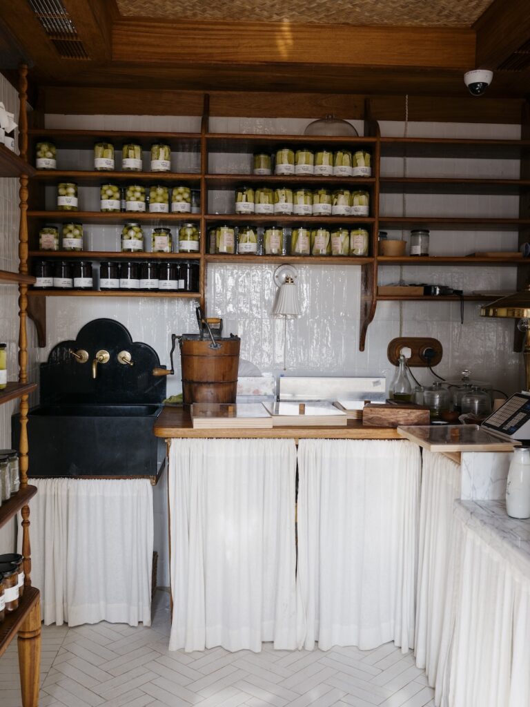 A cozy vintage kitchen with white tile walls, shelves lined with jars of preserved vegetables, a black double sink, a wooden bucket, and counters covered with white curtains. Natural light from the liwan fills the space.