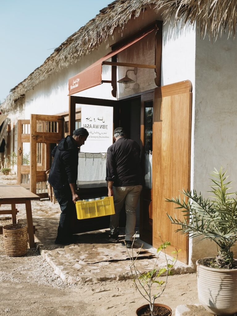 Two people carry a yellow plastic crate into a rustic building with a thatched roof, wooden doors, and a welcoming liwan. Potted plants and outdoor seating are visible around the entrance on this sunny day.