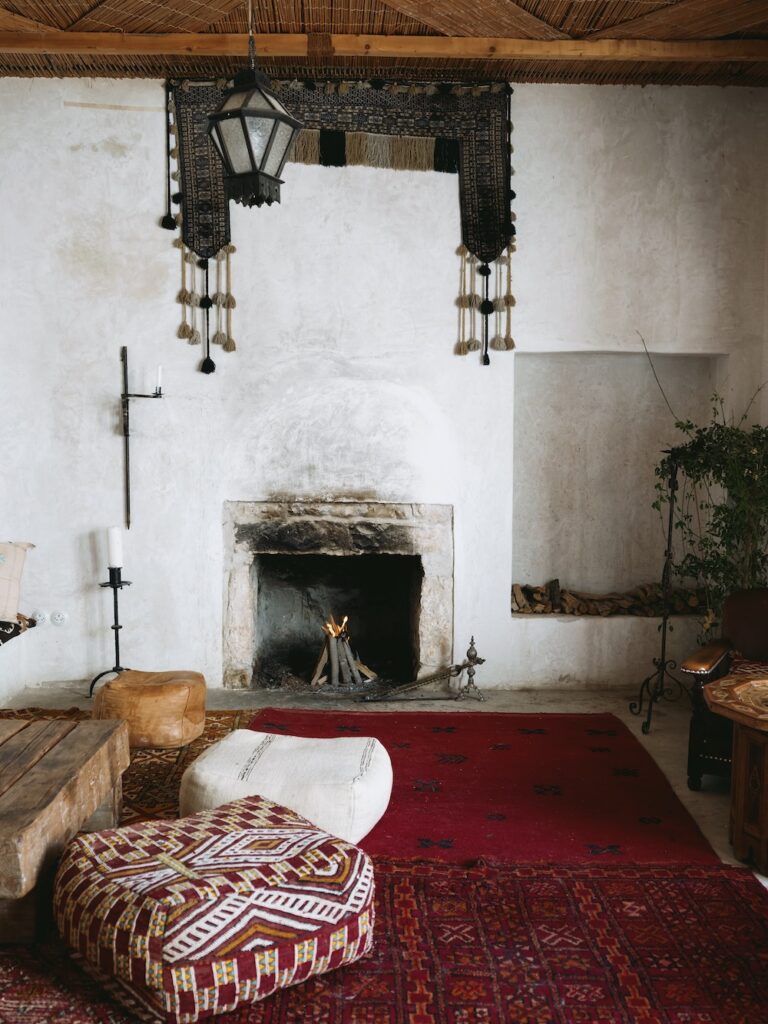 Cozy living room with a white plaster fireplace, a red patterned rug, floor cushions, a wooden bench, and hanging lantern evokes liwan charm; wall art above the fireplace and firewood stacked in a niche add to the warm, rustic decor.