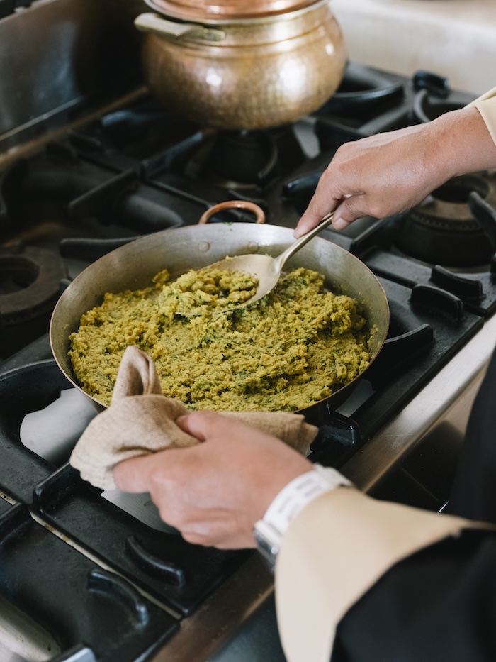A person stirs a pan of greenish, thick food on a stove with a spoon, holding the pan handle with a cloth for safety. A copper pot is visible in the background, adding charm to this cozy liwan kitchen scene.