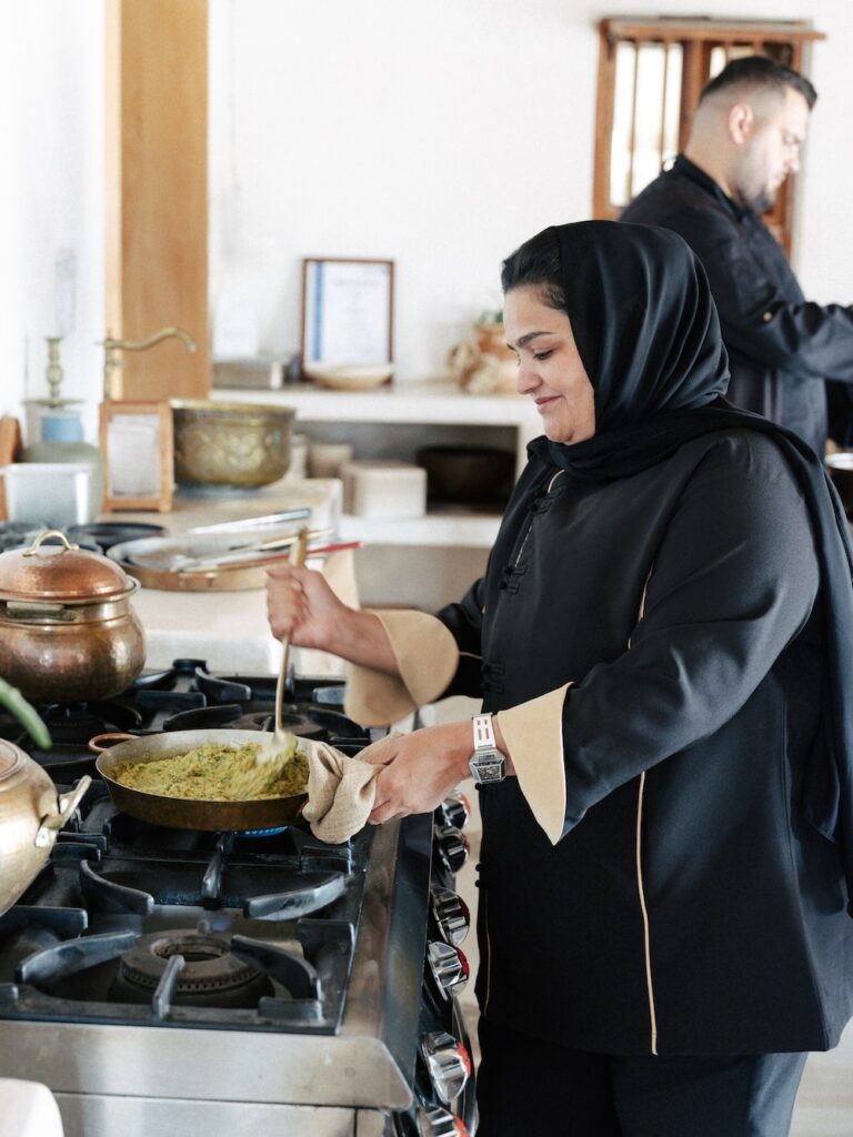 A woman in a black headscarf and clothing stirs food in a pan on the stove of a bright kitchen, while a man works in the liwan area in the background.