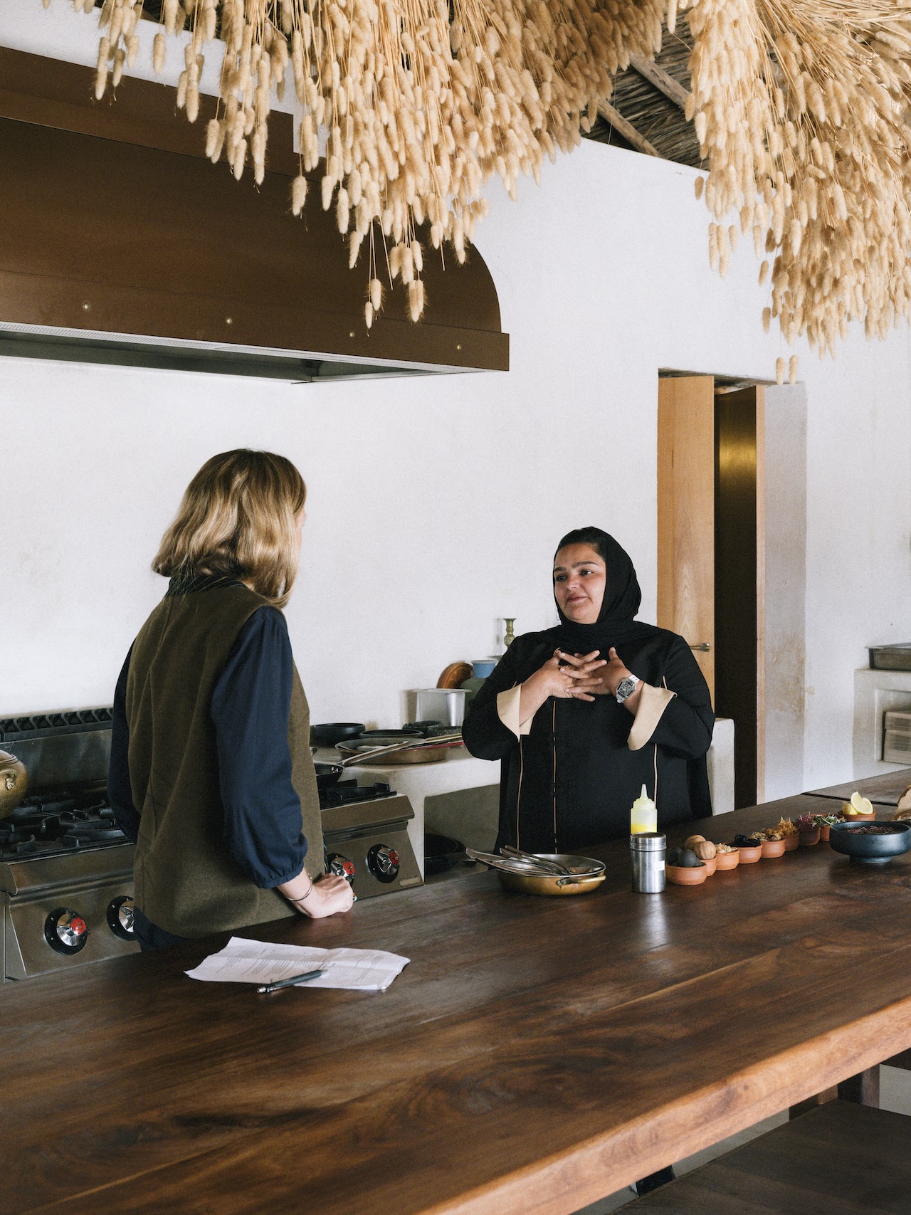 Two women are talking in a modern kitchen with a wooden counter, reminiscent of a liwan. One, wearing a headscarf, gestures with her hands while the other stands across from her. Dried plants hang from the ceiling above them.