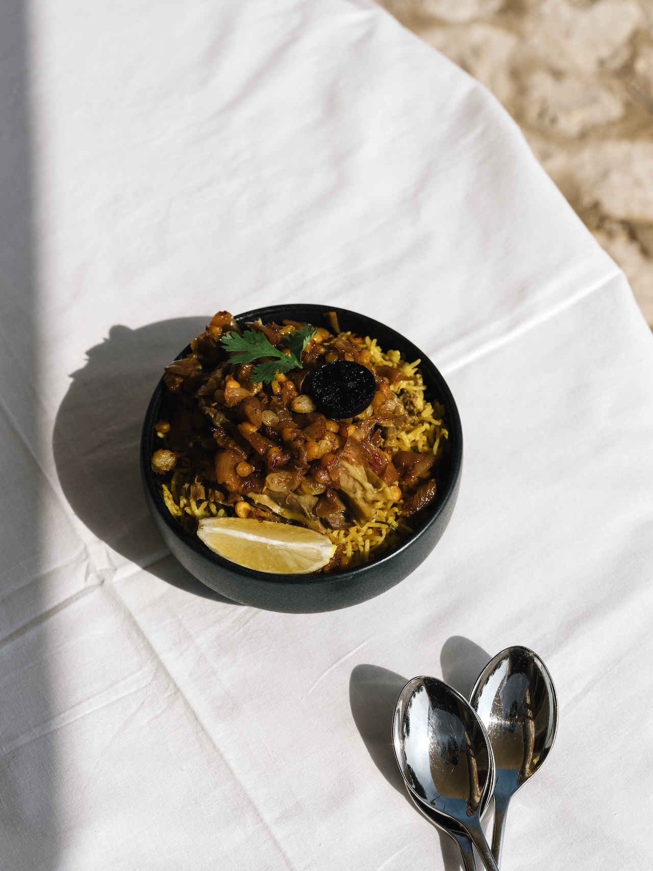 A bowl of yellow rice topped with stewed vegetables, a lemon wedge, and a garnish sits on a white tablecloth beside three silver spoons at Liwan. Sunlight casts soft shadows across the inviting scene.