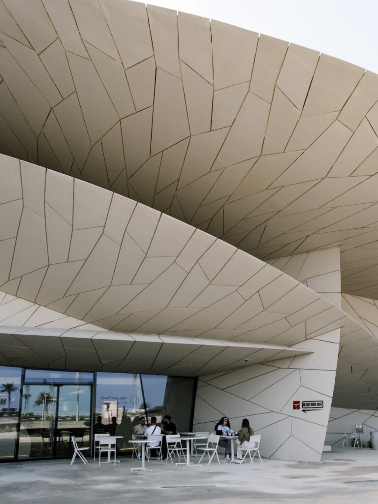 People sit at white tables outside a modern building with large, overlapping, beige panels resembling petals or scales. The geometric, futuristic architecture features glass doors and a clean liwan area in front.