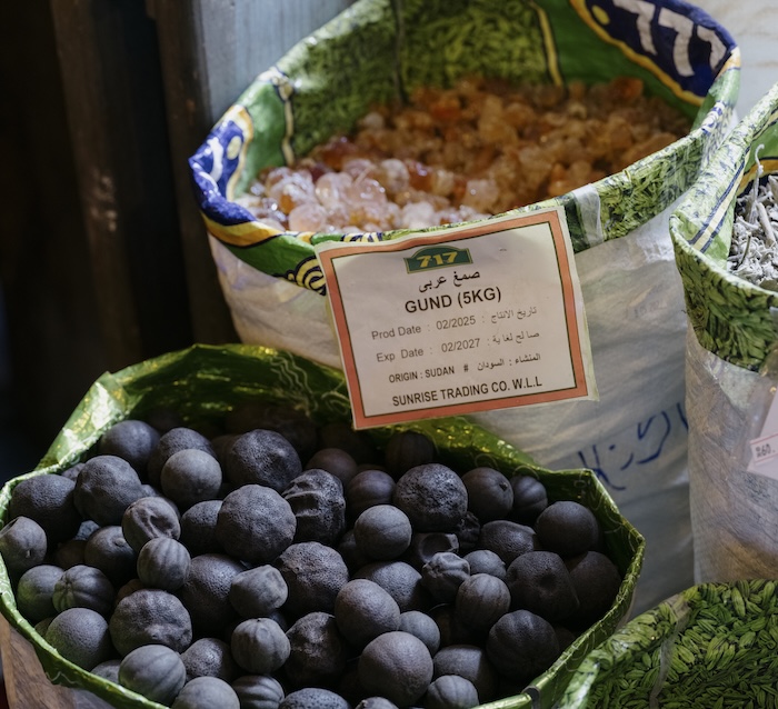 A bag of dried black limes and a bag of gund (gum arabic) from liwan are displayed with labels showing weight, product name, origin (Sudan), and company details at a market.