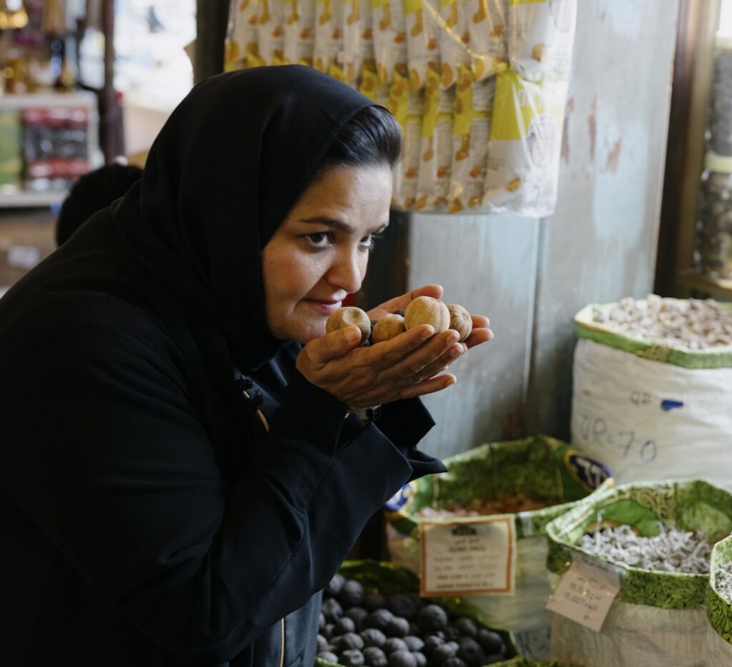 A woman in a black headscarf holds and smells dried goods at a liwan market stall, surrounded by bags of nuts and seeds.