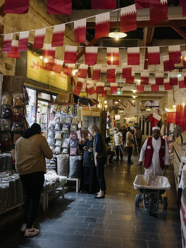 A bustling indoor liwan market with people shopping, Qatar flags overhead, stalls lined with goods, and a man in uniform pushing a wheelbarrow along the stone-paved walkway.