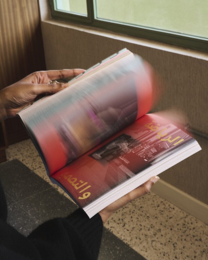 A person wearing a dark sleeve flips through a colorful book with blurred, fast-moving pages. Arabic text and a grayscale photo of a person appear on one page, while soft light enters from a liwan window indoors.