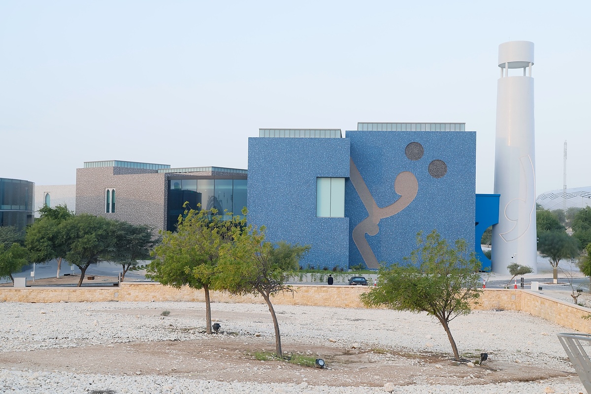 A modern building with a blue mosaic façade featuring abstract designs, large windows, and a tall white tower—home to the Lawh Wa Qalam Museum—stands behind a few trees and a rocky foreground under a clear sky.