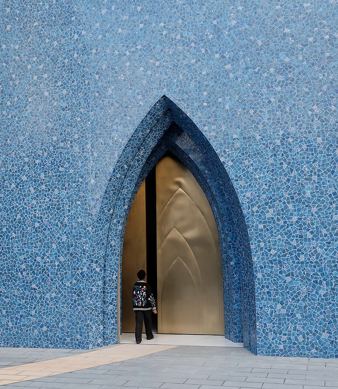 A person stands at the entrance of the Lawh Wa Qalam Museum, framed by a tall, arched doorway set in a blue mosaic-tiled wall and golden doors partially open. The person carries a bag and wears a patterned jacket.