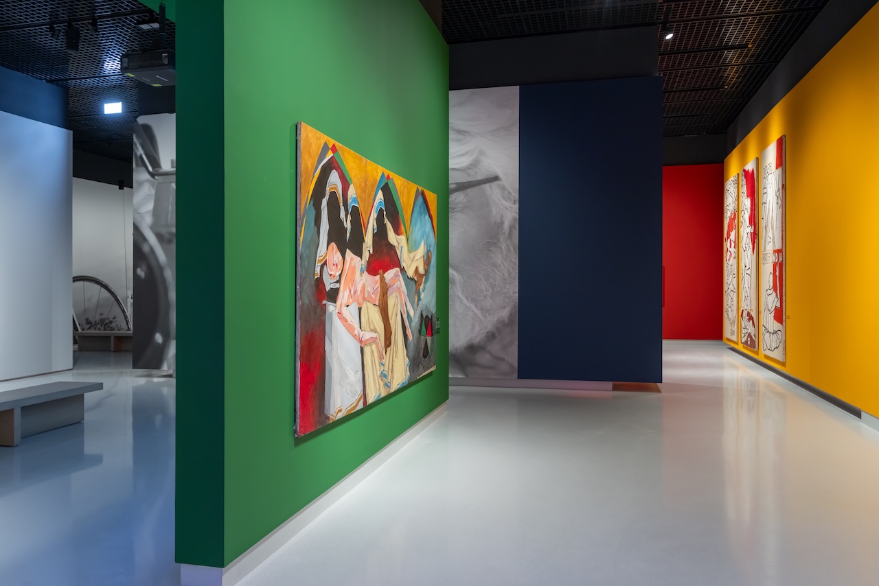 A modern art gallery, the Lawh Wa Qalam Museum, features colorful walls displaying large abstract paintings. One green wall showcases vibrant artwork, while dark blue, red, and yellow walls exhibit expressive contemporary pieces.