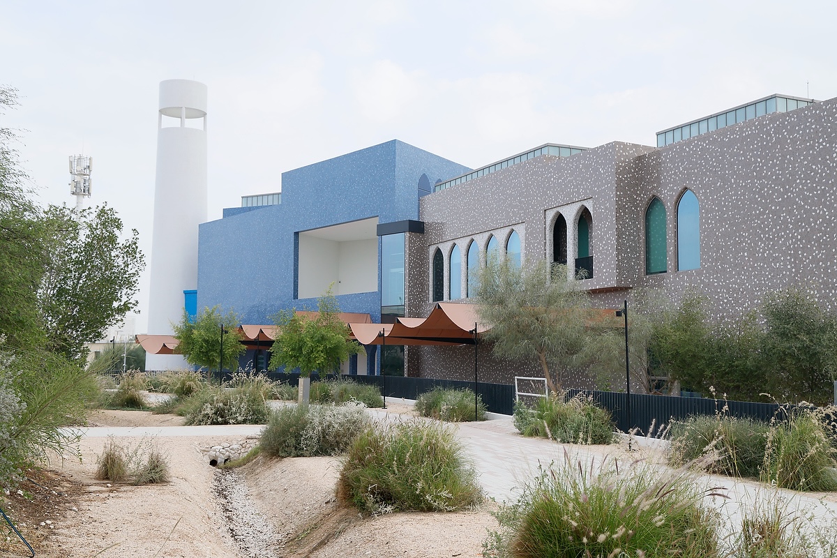 Modern building with blue and gray geometric facades, arched windows, and a tall white tower in the background—home to the Lawh Wa Qalam Museum—surrounded by trees, plants, and a landscaped walkway under a partly cloudy sky.