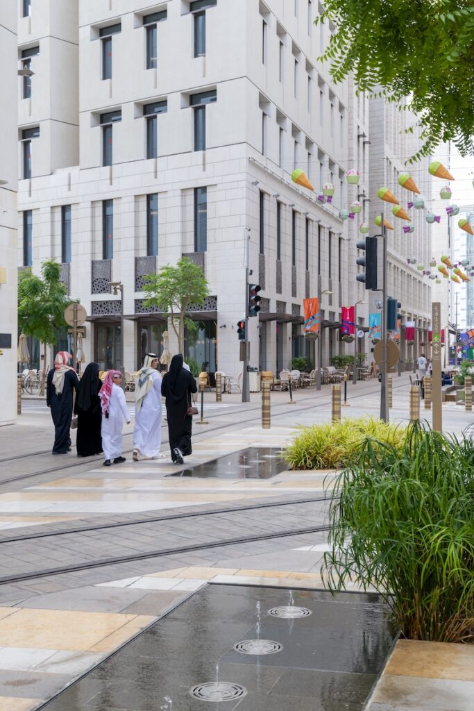 A group of people in traditional Middle Eastern attire walk along a modern, clean city street in Msheireb, lined with trees, colorful lanterns, and tall white buildings on a cloudy day.