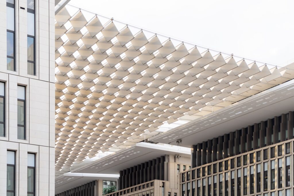 A modern architectural structure in Msheireb features a geometric, lattice-like canopy spanning between tall office buildings with vertical window designs. The canopy creates an intricate shadow pattern. The sky is overcast.