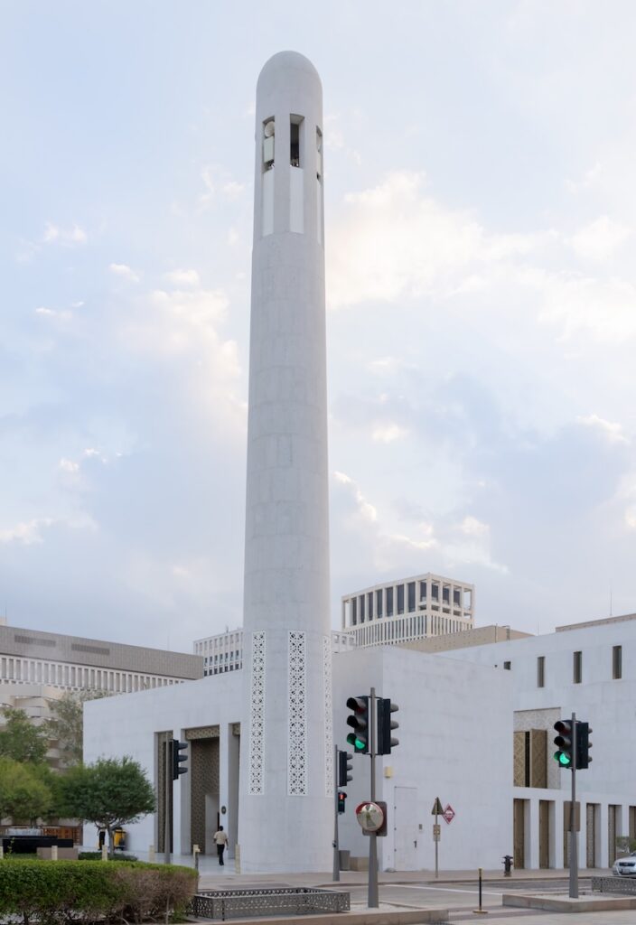 A tall, slender white minaret stands beside a modern Msheireb mosque building, with geometric patterns on the walls. Traffic lights and greenery appear in the foreground under a cloudy sky.
