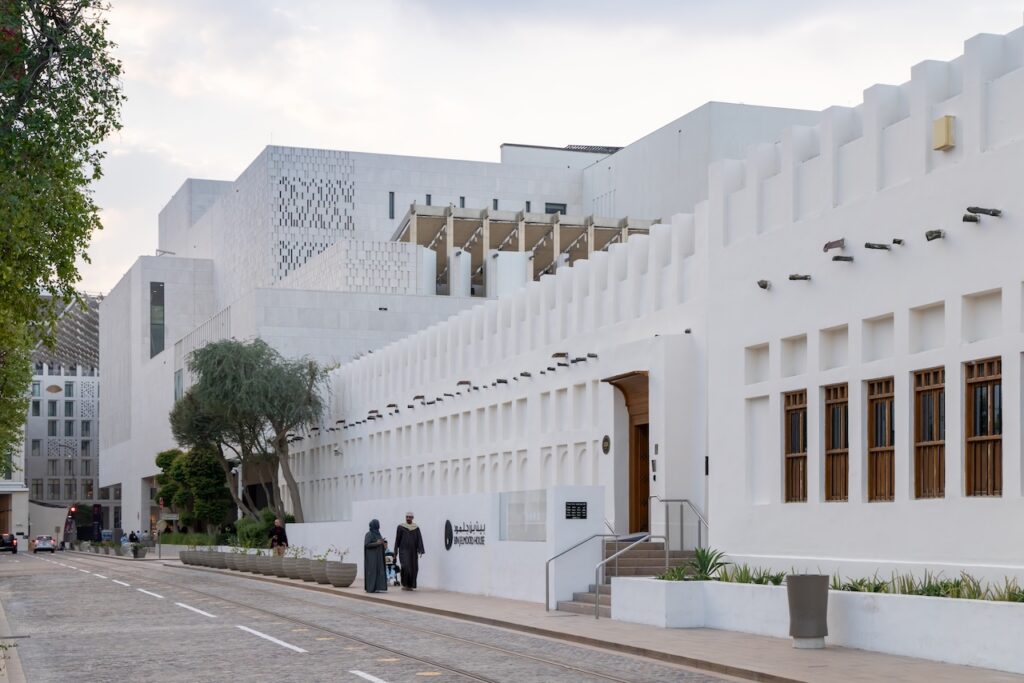A modern white building with geometric designs and wooden windows stands along a cobblestone street in Msheireb. People walk and sit nearby, while trees and plants line the sidewalk under a partly cloudy sky.