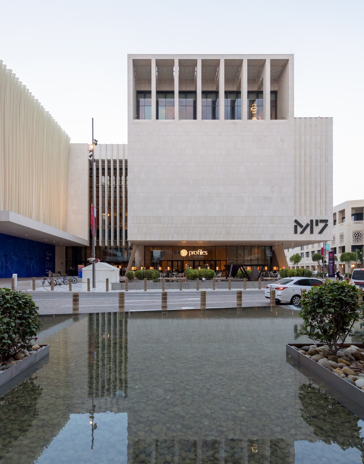 A modern white stone building in Msheireb with large windows and the sign M7 stands behind a shallow reflecting pool with plants. The ground floor features shops and people walking nearby, while cars are parked along the street.