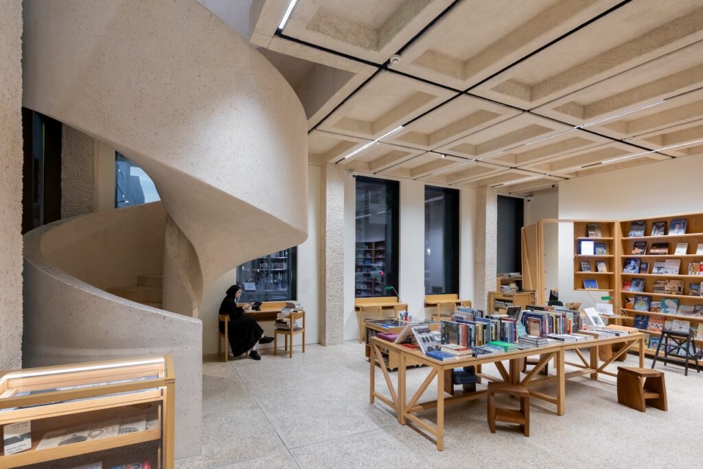 A modern Msheireb library interior with a concrete spiral staircase, wooden tables displaying books, tall bookshelves, and large windows. A person in dark clothing sits reading at a small table near the staircase.