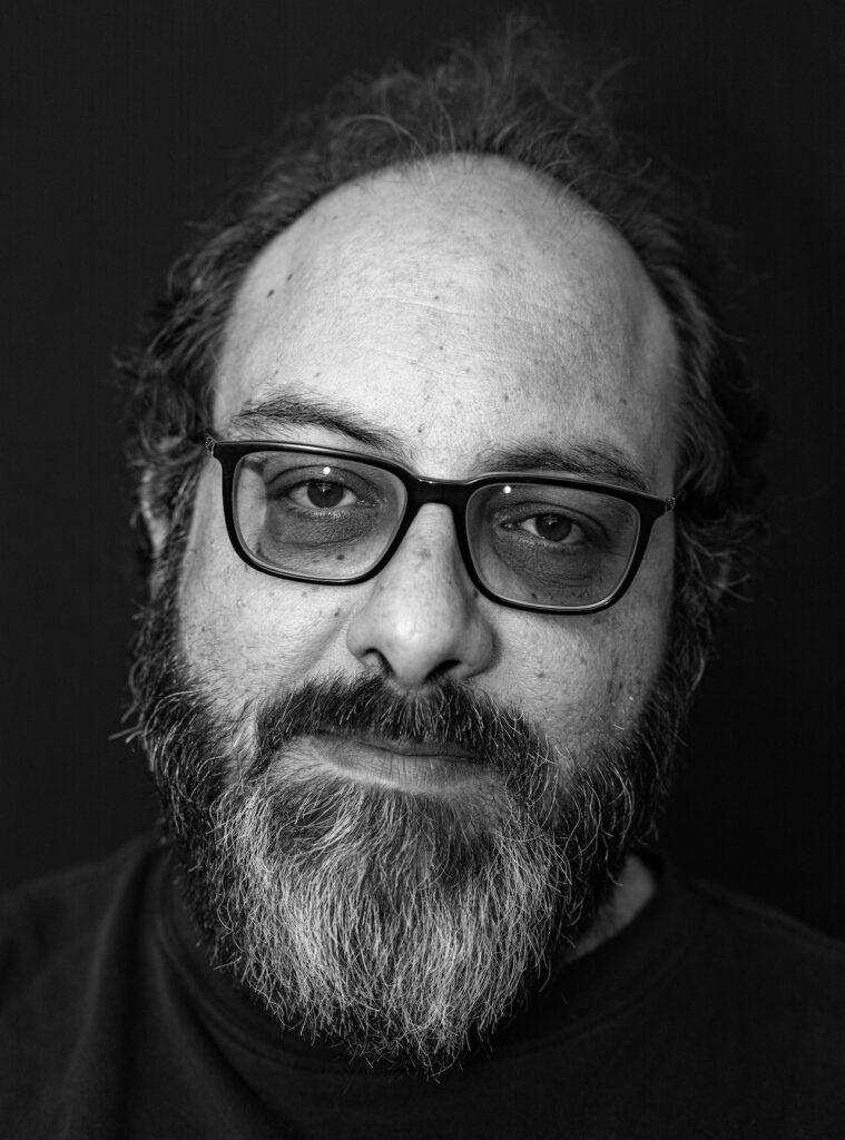 A man with glasses, a full beard, and mustache, looking directly at the camera with a neutral expression. The plain, dark background and black-and-white style evoke portraits of artists like Rirkrit Tiravanija.