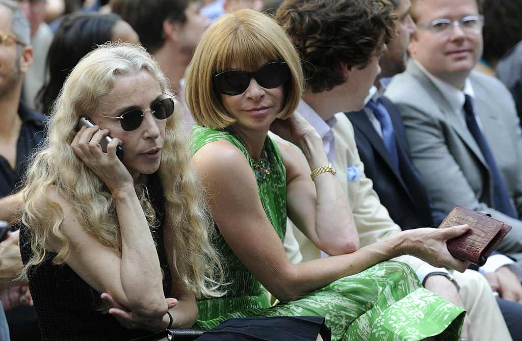Two women wearing sunglasses sit in the front row at the Franca Fund Gala. One has long blonde hair and is talking on a phone, while the other, in a green dress with a short bob haircut, enjoys the outdoor event surrounded by other guests.