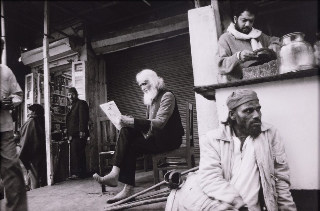 A bearded man sits on a chair reading a newspaper on a busy street near the Lawh Wa Qalam Museum. Another man stands at a counter behind him, preparing food or drink. A third man with crutches sits on the ground, looking away in this lively, candid scene.