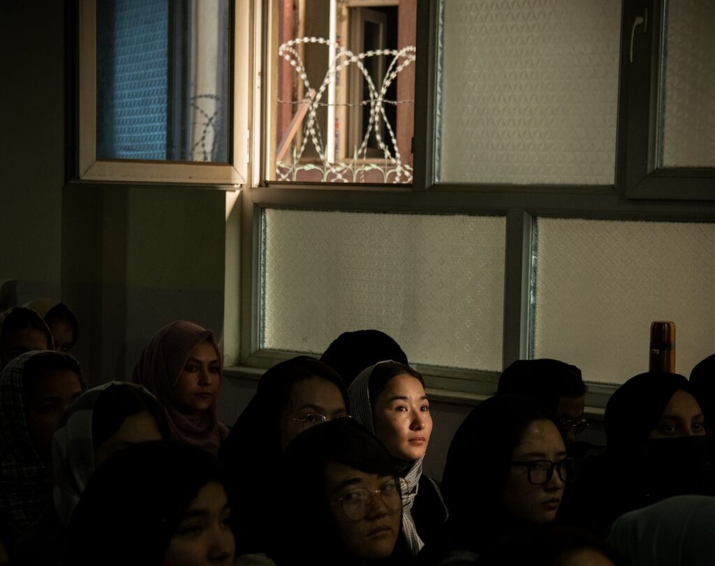 A group of women, some wearing headscarves, sit closely together in a dimly lit room. Sunlight—an empire of light—shines on one woman’s face, drawing attention to her. Behind them, a window with barbed wire outside is partially open.