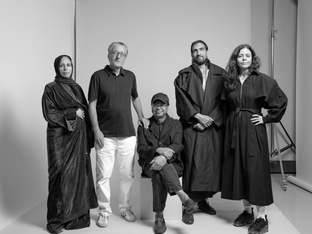 A black and white photo of five people posing in a studio echoes the style of Rirkrit Tiravanija. Four stand while one sits, all in dark, loose-fitting outfits—two women in dresses and three men in jackets or casual clothing.
