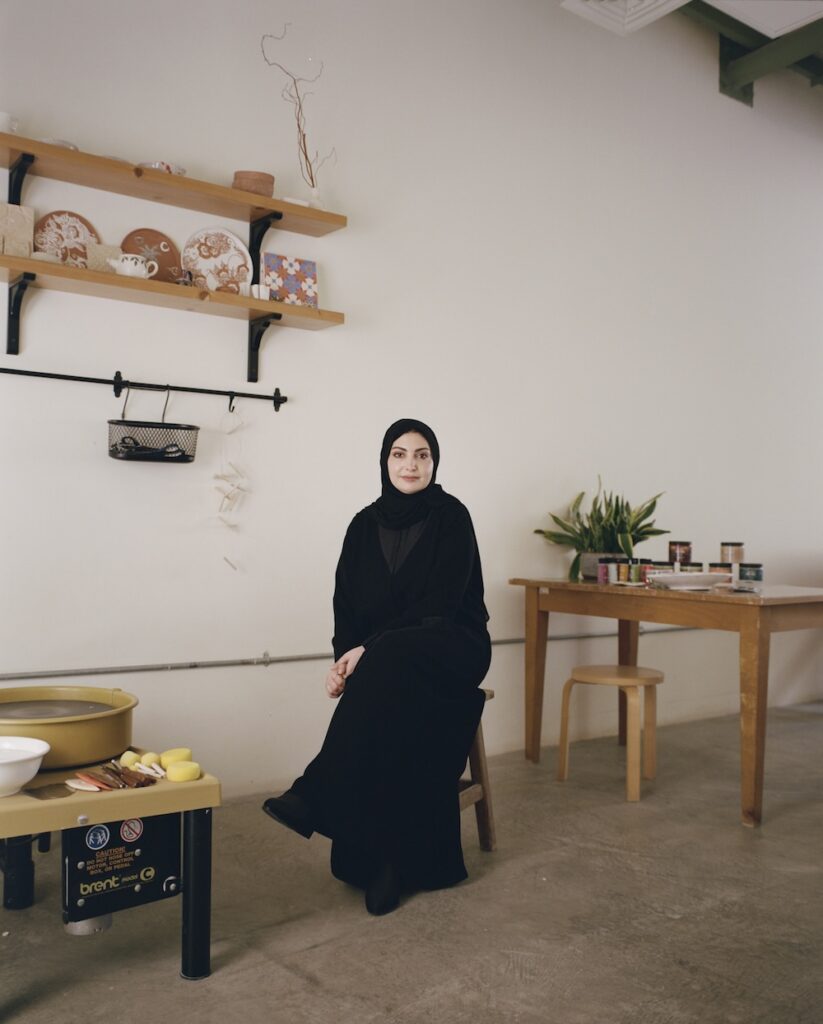 A woman in a black abaya and hijab sits on a stool in a minimalist pottery studio inspired by liwan design, surrounded by shelves with ceramic plates, a table with art supplies, and a potted plant on the floor.