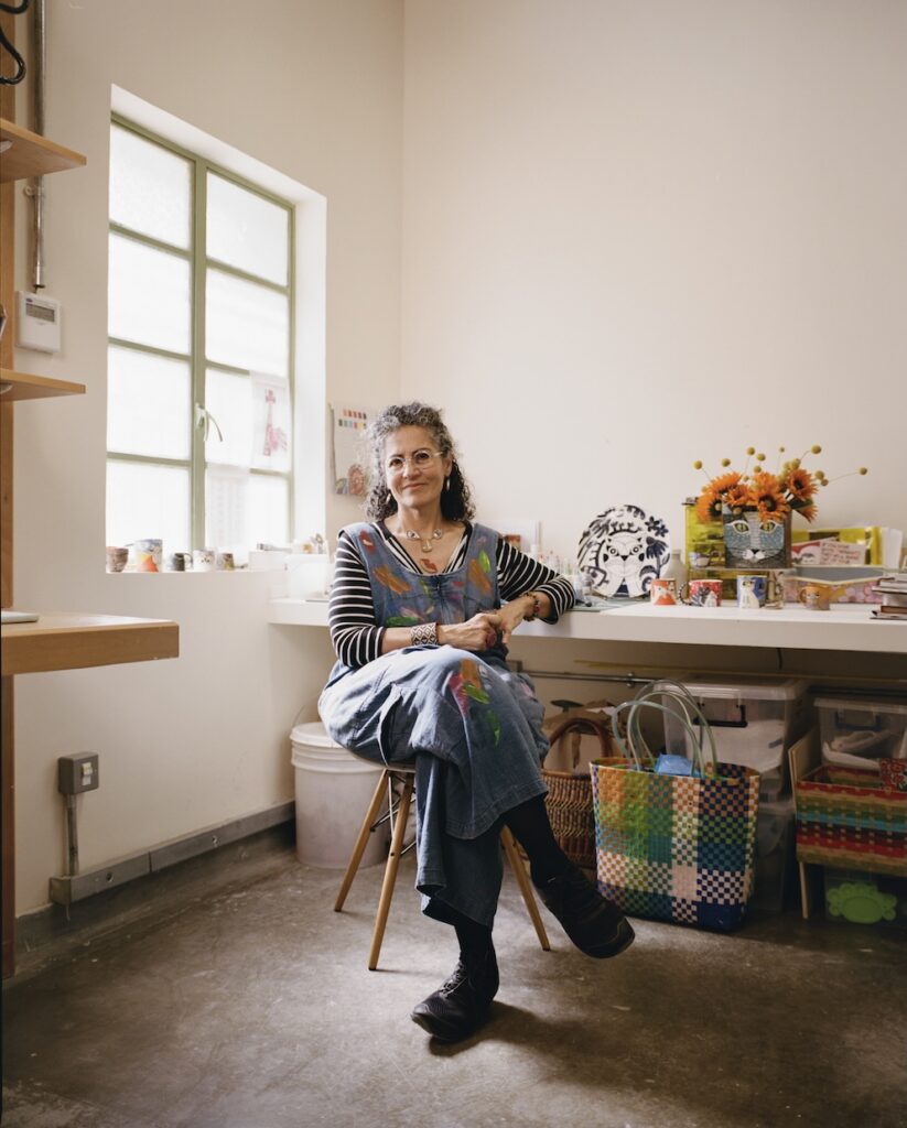 A woman with curly hair and glasses, dressed in a striped shirt and denim overalls, sits on a chair in a bright liwan studio filled with art supplies, colorful baskets, and flowers on a table beside her.