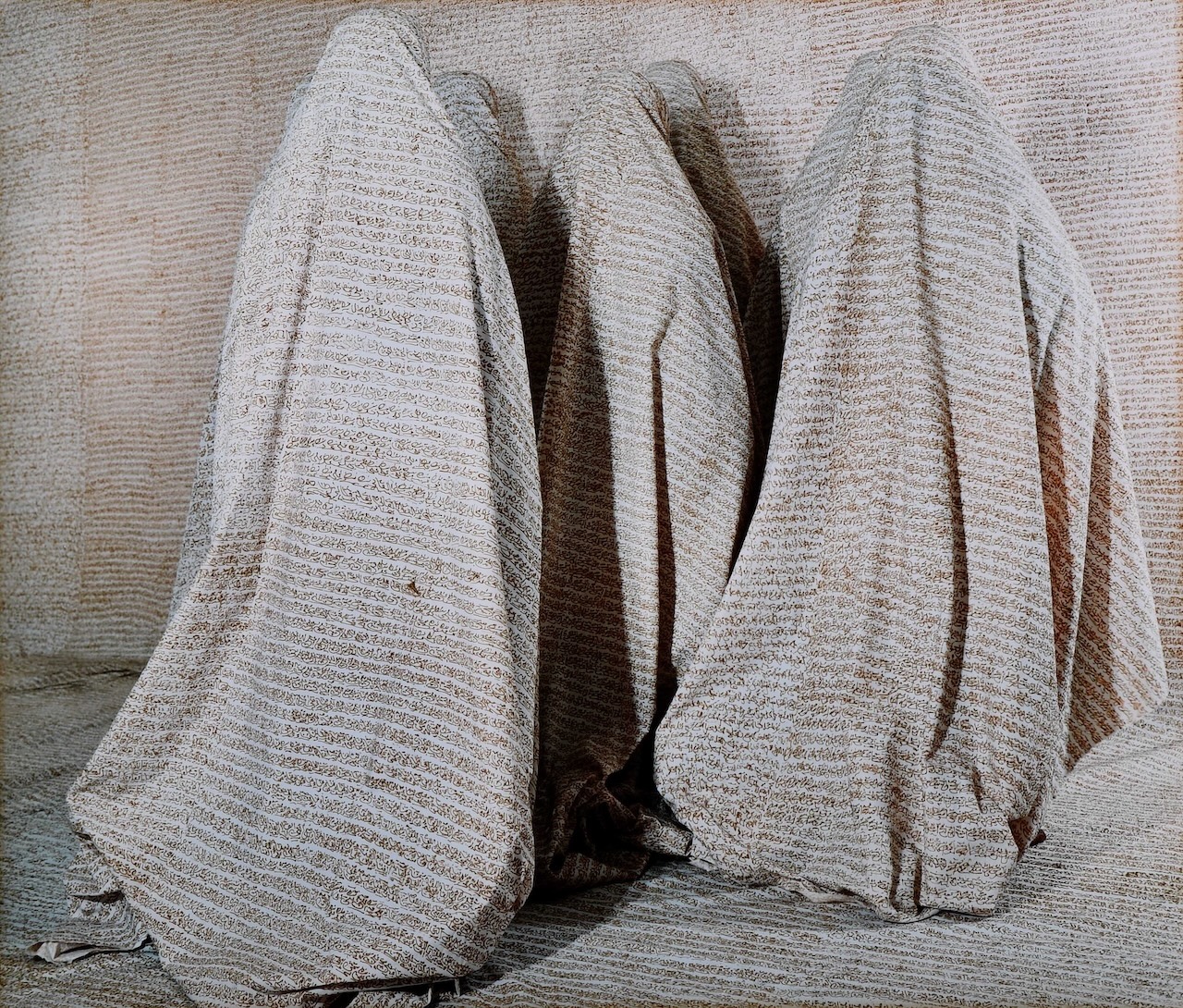 Three human portraits are entirely covered with striped, textured beige fabric, blending into a similarly patterned background and creating a mysterious, camouflaged effect. Their shapes are only visible beneath the draped cloth.