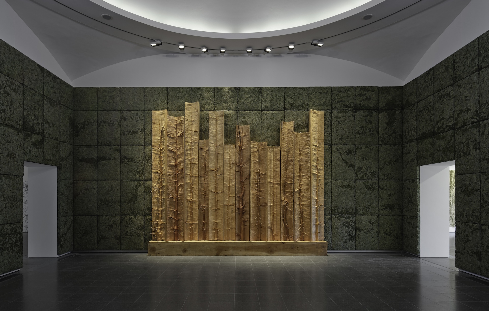 A modern art installation featuring tall, vertical wooden beams of varying heights displayed against a textured, green wall in a well-lit, spacious gallery room with a curved ceiling.