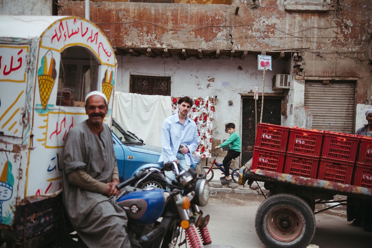 A man sits on a motorcycle beside a colorful ice cream cart on a busy street. Behind him, a man walks and a child runs past red crates, while old, weathered buildings echo the vibrant energy of saint levant in the background.