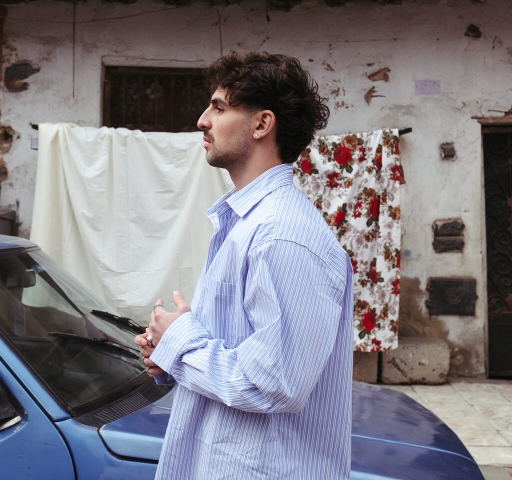 A man in a blue striped shirt stands beside a blue car, looking to the side like Saint Levant. Behind him, a white and a floral sheet hang against an old, weathered building.