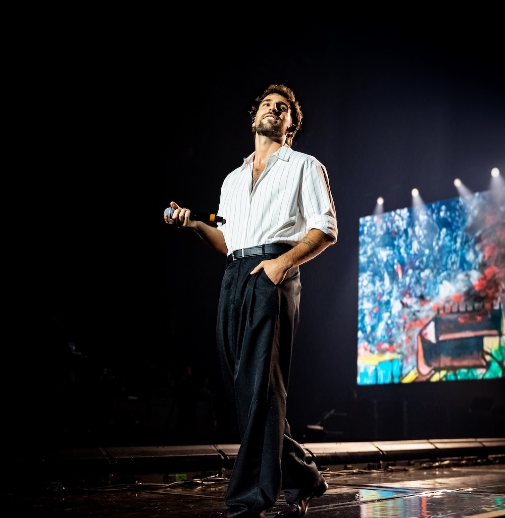 A man in a white shirt and dark pants stands on stage holding a microphone, with colorful abstract visuals displayed on a large screen behind him, as stage lights shine—channeling the dynamic energy of Saint Levant.