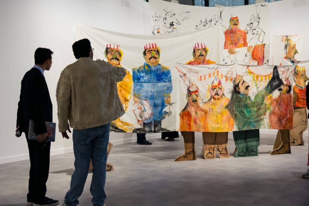 Two people view an art installation at ArtBasel Qatar featuring painted fabrics hung up, each depicting colorful figures in uniforms and hats; some hold banners, with cartoonish figures drawn on the wall behind.