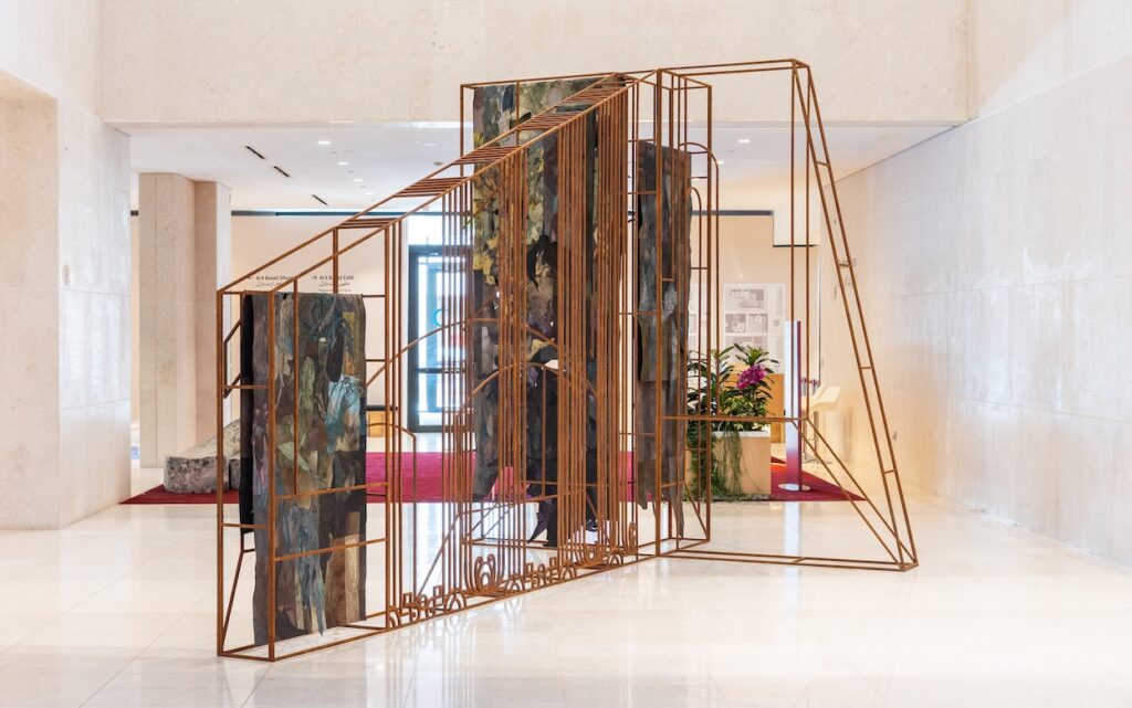 A geometric metal sculpture with vertical bars and rectangular panels featuring abstract art, inspired by ArtBasel Qatar, stands in a marble-floored, modern lobby with beige walls and a potted plant in the background.