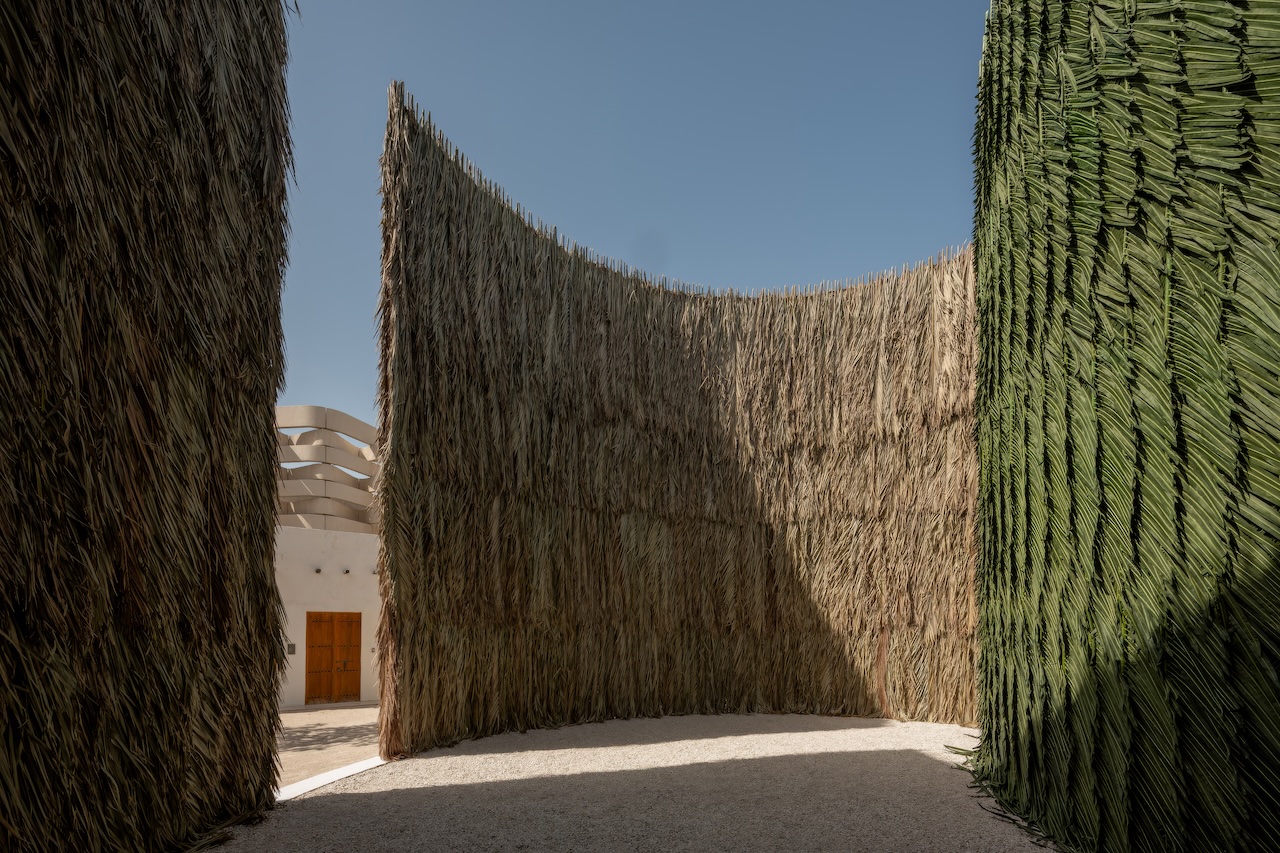 Tall, curved walls made of tightly woven dried palm fronds form an outdoor enclosure under a clear blue sky at ArtBasel Qatar, with a white building and wooden door visible in the background.