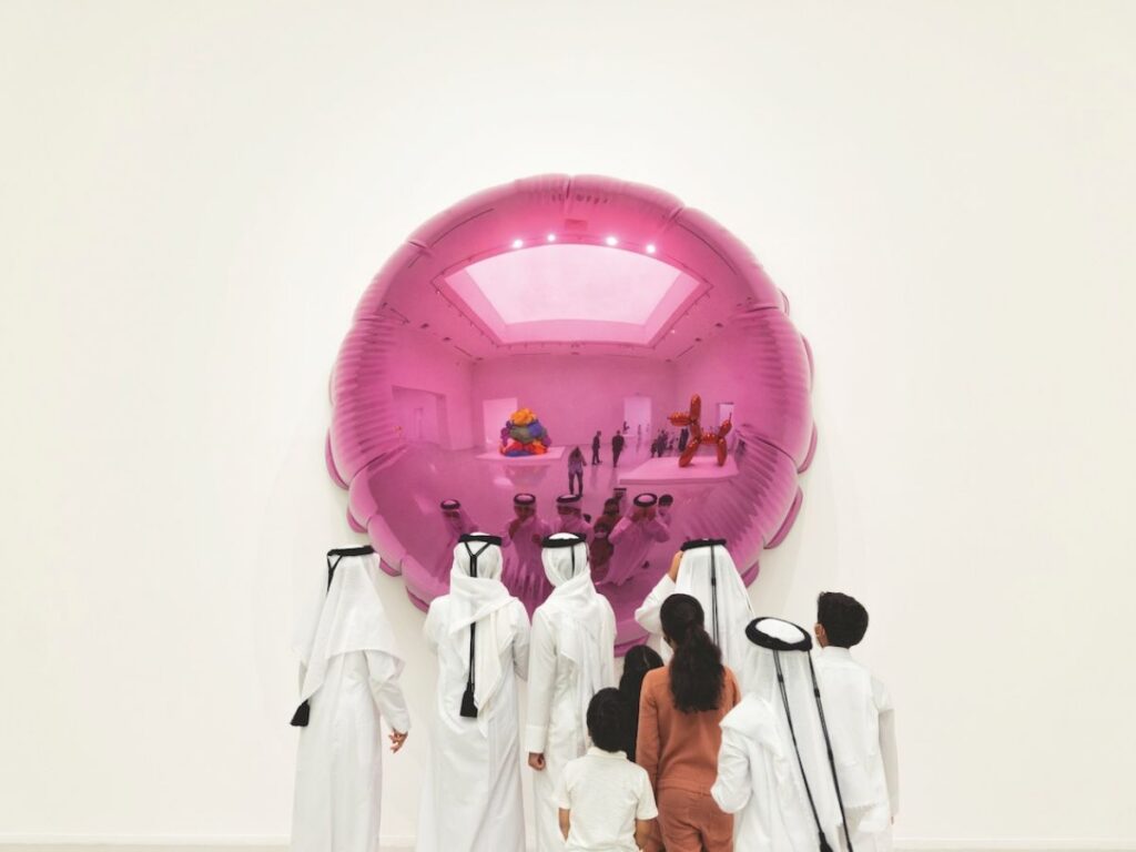 A group of people in traditional Middle Eastern clothing stand facing a large, pink, reflective wall sculpture at a Qatar Creates event that mirrors their image and the gallery space around them.