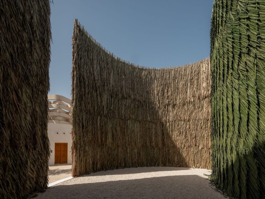 Tall, curved walls made of dried palm fronds enclose an open outdoor space under a clear blue sky—an architectural gem reminiscent of installations seen at ArtBasel Qatar. A wooden double door stands in the light-colored building in the background.
