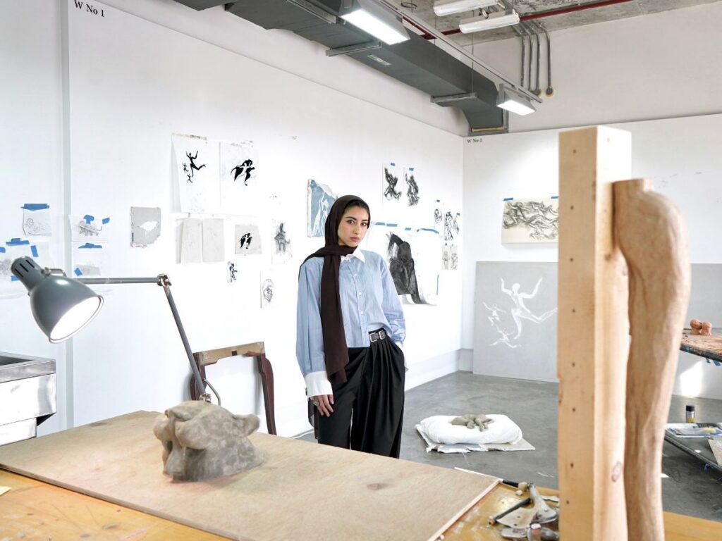 Alla Abdunabi stands in an art studio surrounded by drawings on the wall, sculptures, and wooden art pieces. She wears a blue shirt, black pants, and a scarf, looking toward the camera with a calm expression.