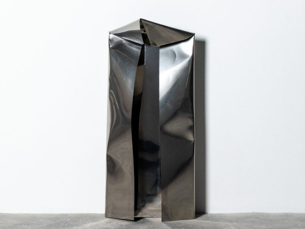 A tall, shiny, metallic sculpture with sharp, angular folds stands against a plain white wall on a gray floor, evoking gulf futurism as it resembles an otherworldly folded sheet of metal.