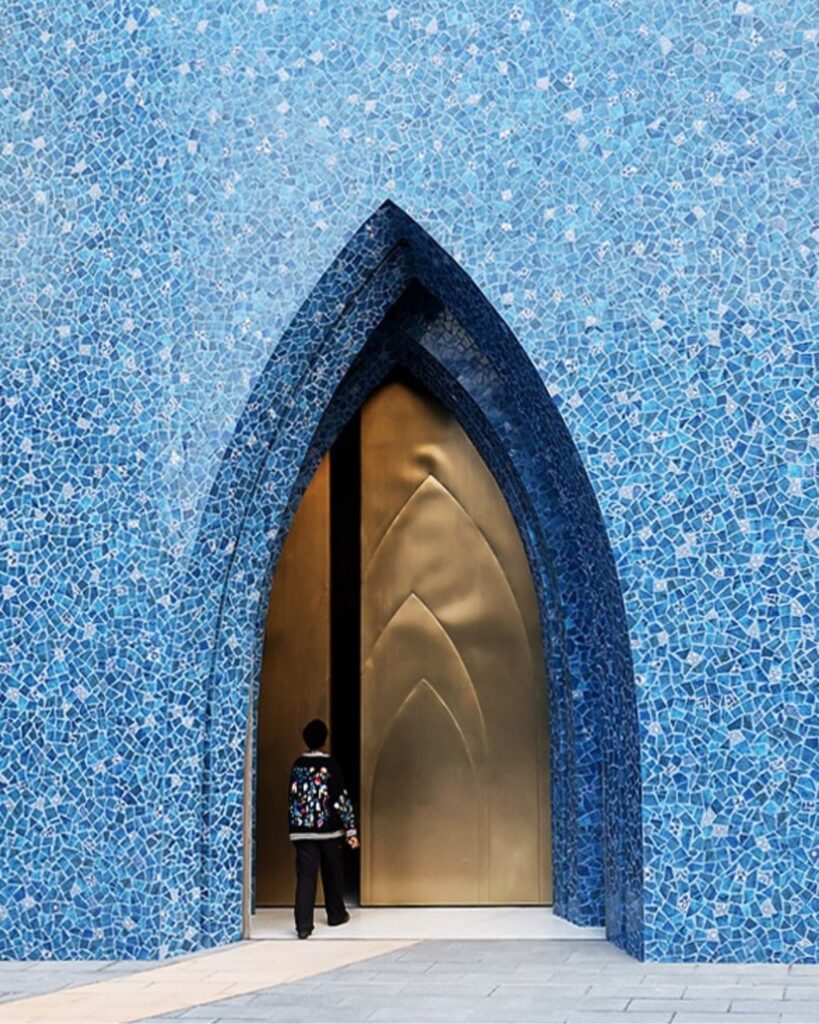 A person stands at the entrance of the Lawh Wa Qalam Museum, framed by blue mosaic tiles and a tall, pointed arch doorway, about to step inside through a golden door.