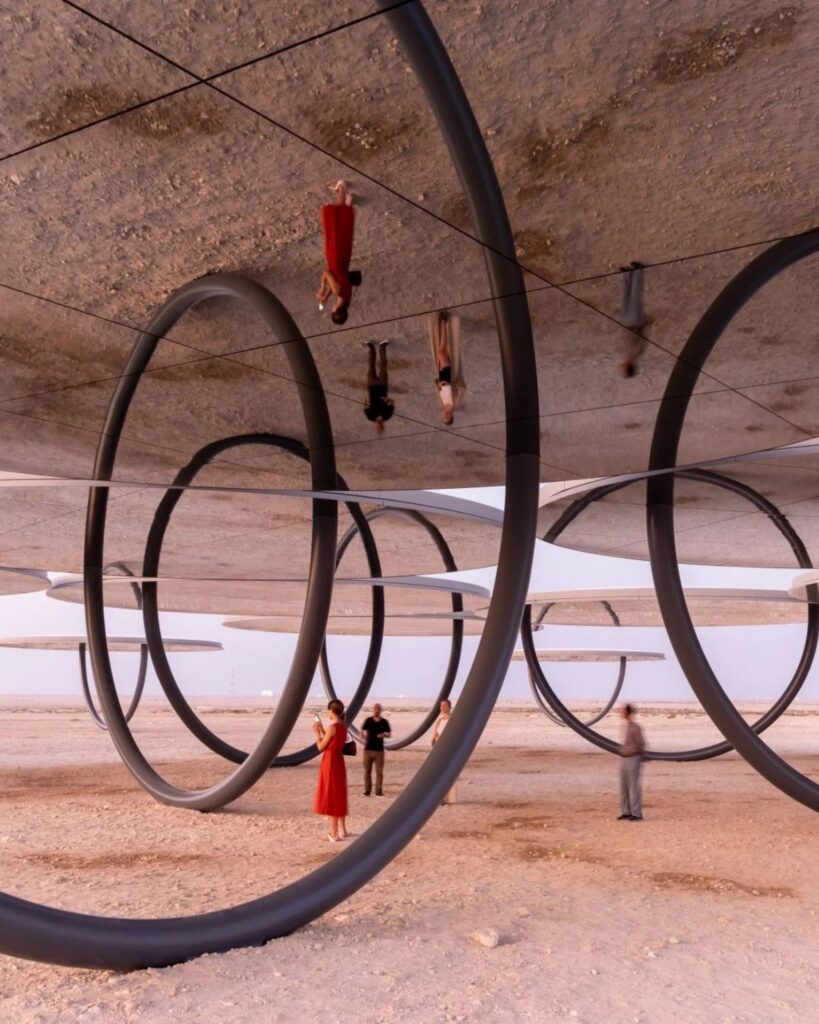 People stand and walk among large circular public art sculptures on a desert landscape, with some of their reflections appearing upside down on mirrored surfaces above them. The sky is clear and the scene is surreal and artistic.