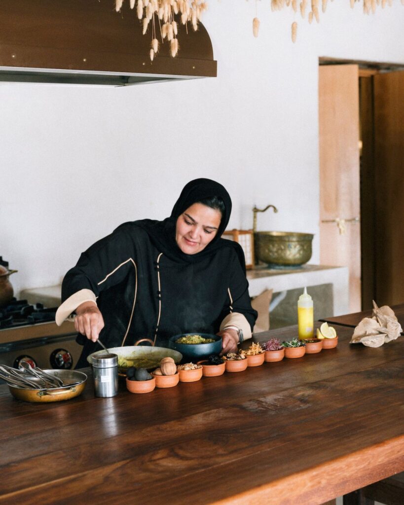 Chef Noof Al Mari, in a black headscarf, cooks at a wooden kitchen counter lined with spice bowls. She stirs food in a pan, with kitchen utensils and a yellow bottle nearby, in a warm and rustic room.