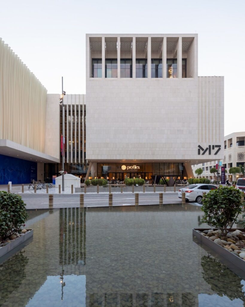 A modern white building with large square windows, labeled M7, stands behind a shallow reflecting pool with clear water and greenery on both sides in Msheireb. Cars and pedestrians are visible near the entrance.