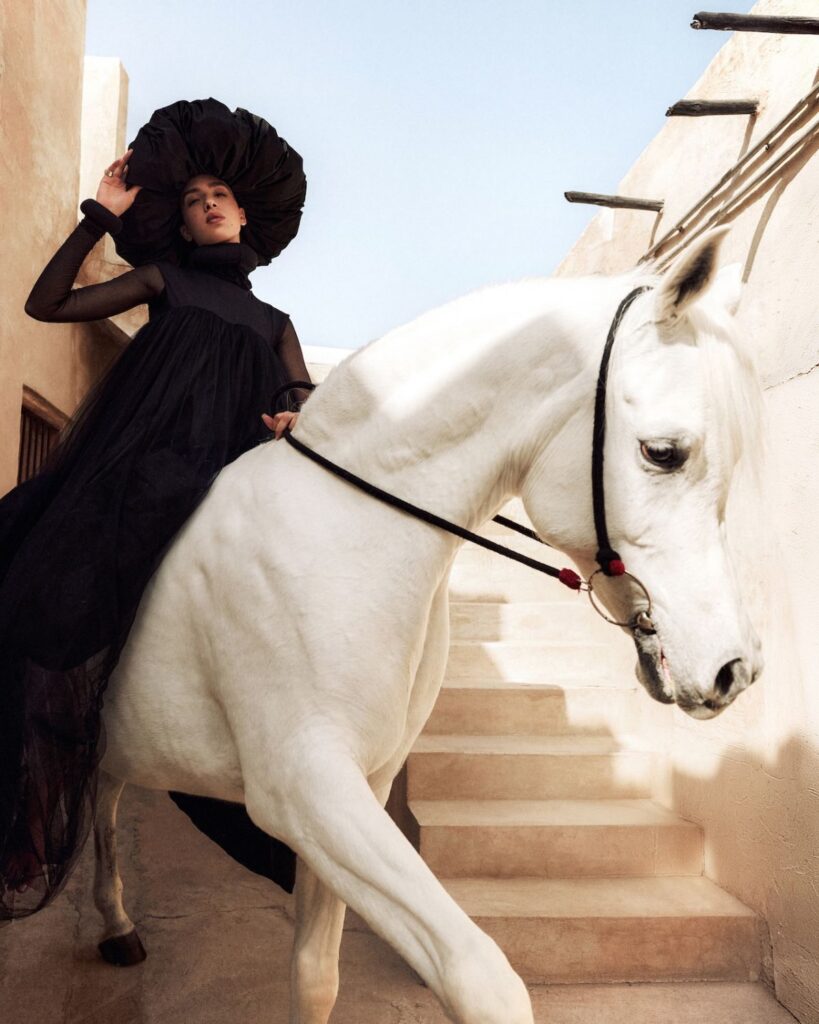 A woman in a dramatic black dress and large hat, evoking the elegance of the Fashion Trust Arabia Prize, poses on a white horse rearing up on a stone staircase against beige walls and a blue sky.