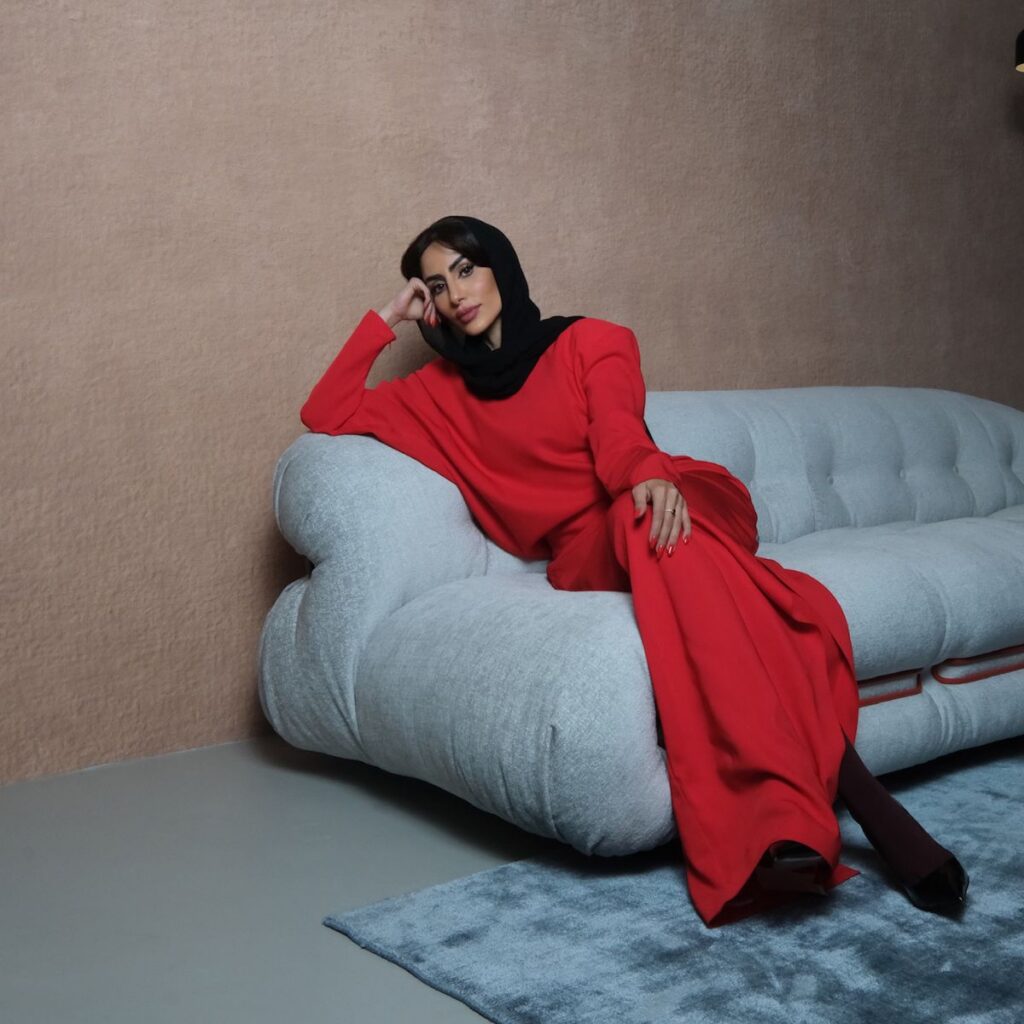 Shaikha Al Sulaiti, wearing a black headscarf and a long red dress, sits relaxed on a light gray sofa against a beige wall, resting her head on her hand. A blue-gray rug is spread on the floor in front of her.