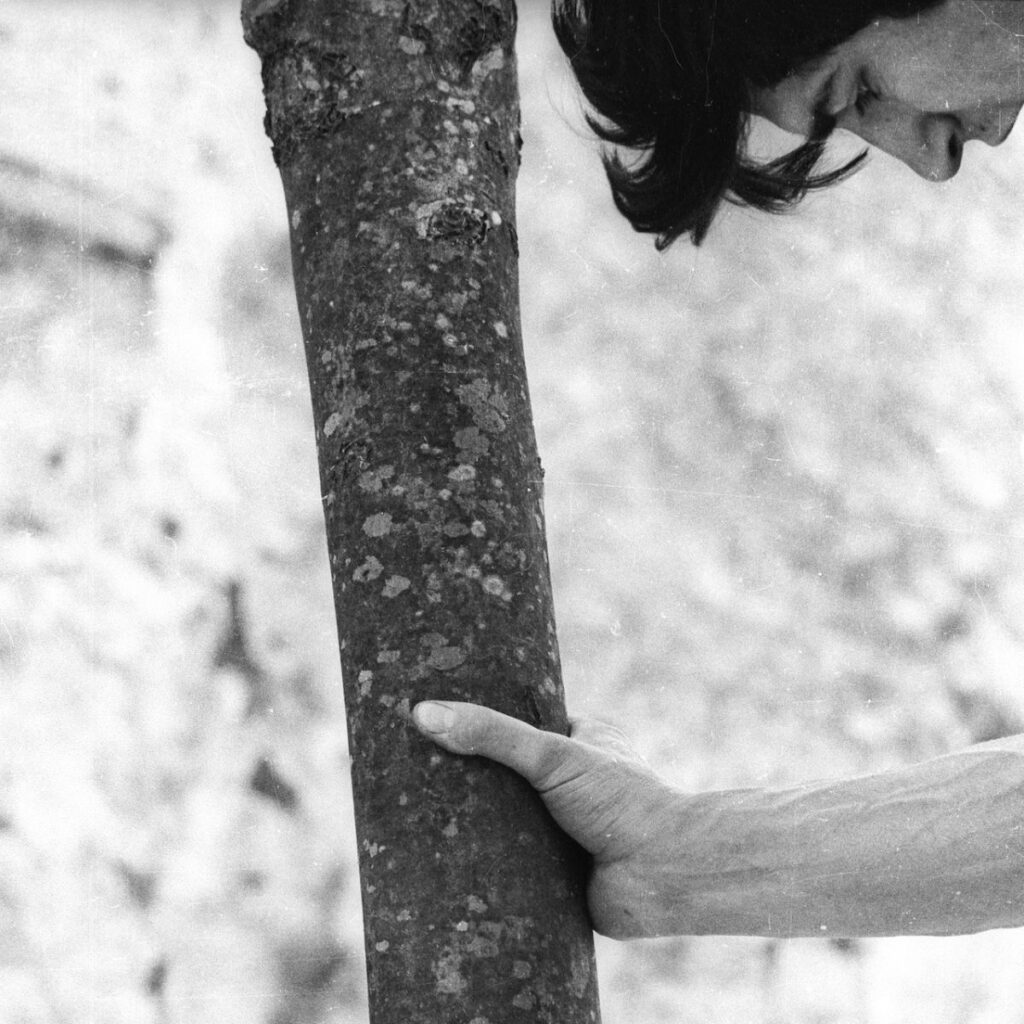 A person tightly grips a tree trunk with one hand, head lowered in deep focus or effort—a moment reminiscent of the contemplative scenes often found in Rirkrit Tiravanija’s work. The scene is rendered in black and white, with the background blurred into light, soft shapes.