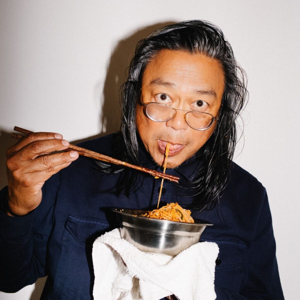 A person with long dark hair and glasses, reminiscent of a Rirkrit Tiravanija artwork, eats noodles from a metal bowl using chopsticks, holding the bowl with a towel against a white background.
